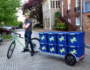 Man on bike standing with arms crossed. Blue bins with Annapolis Compost logo piled up on bike trailer.